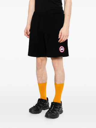Person wearing black shorts with a logo by Canada Goose, orange socks, and black shoes on a white background