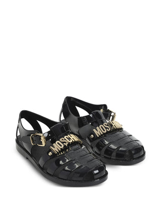 Moschino black logo buckle sandals with gold-tone hardware, open toe, and flat sole. A graphic warm-weather staple, available at our Lisbon store.