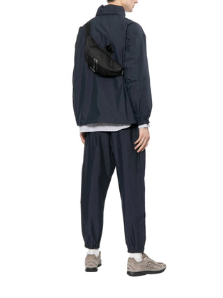 Person wearing a dark jacket and pants with a black bag over one shoulder 