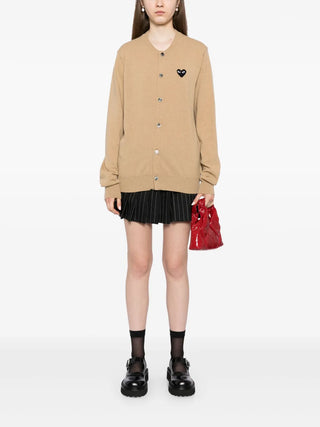 Person wearing a camel brown cardigan with a black heart emblem, black pleated skirt, and black shoes 