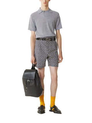 Person wearing a gray polo shirt, patterned shorts, yellow socks, and black shoes, holding a black backpack 